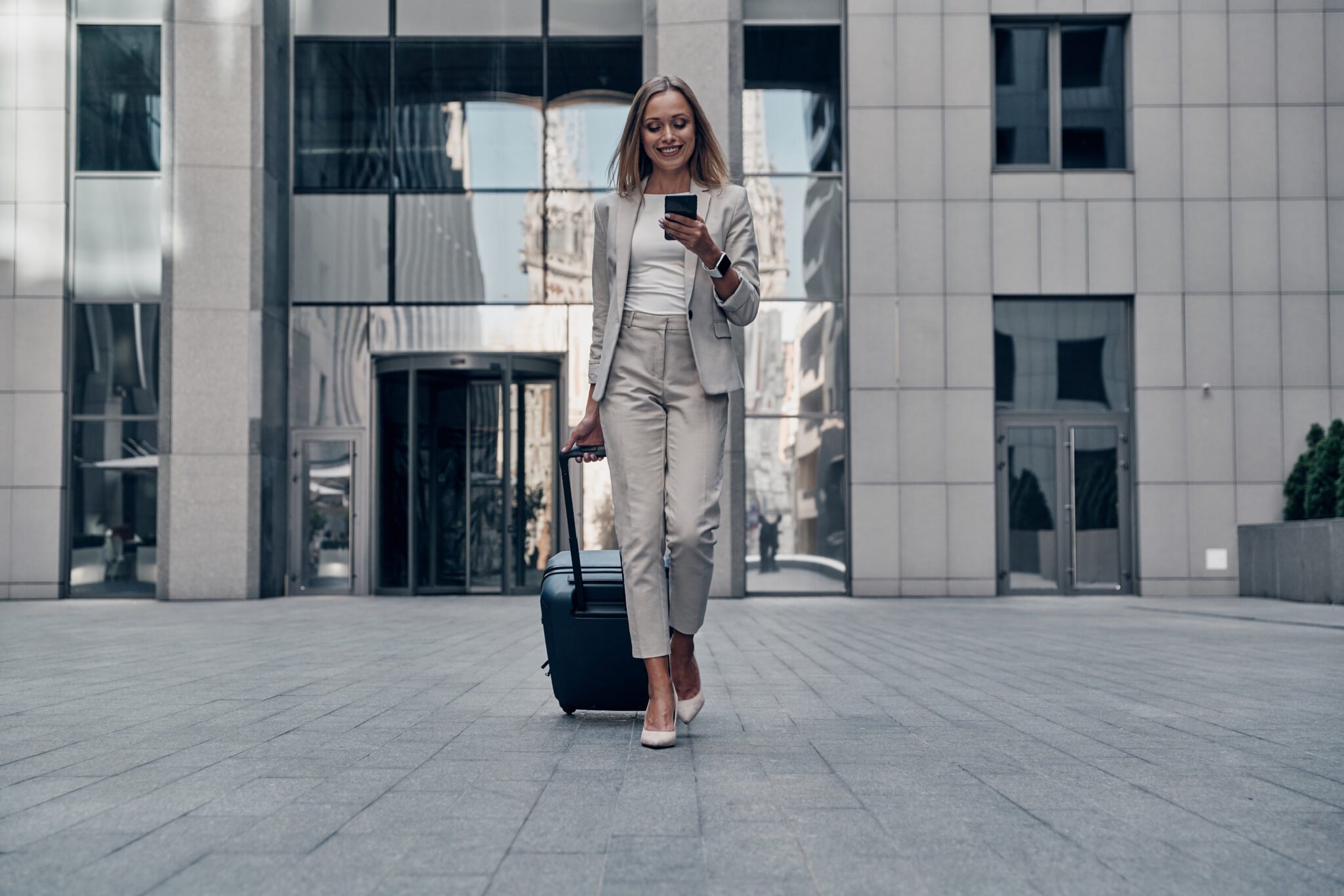 Always ready to help. Full length of young woman in suit pulling luggage and smiling while walking outdoors